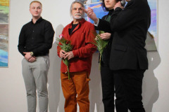 Przy ścianie z obrazem stoją w rzędzie bohaterowie wystawy i kurator Robert Brzęcki z mikrofonem.