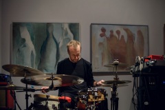 Krzysztof Szmańda gra na perkusji. Za jego plecami dwa duże obrazy zawieszone na białej ścianie.