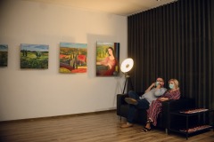 Dwoje ludzi siedzi na czarnej kanapie. Na ścianie wiszą cztery kolorowe obrazy. W rogu stoi zapalona lampa.
