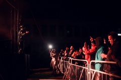 Po lewej stronie wokalista stoi przy krawędzi sceny. Po prawej publiczność zgromadzona przy barierkach.
