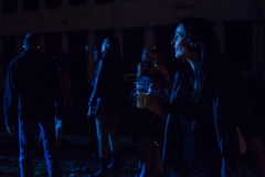 Uśmiechnięta kobieta z czarnymi słuchawkami na uszach, trzyma kubek piwo. Spogląda w stronę sceny. Jej twarz rozjaśnia białe światło.