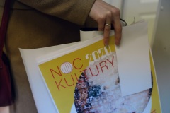 Tegoroczny plakat Noc Kultury 2020 trzymany przez kobietę. Widoczny fragment dłoni i brązowego płaszcza.