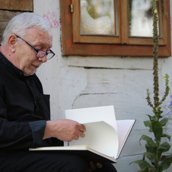 Włodek Sypniewski siedzi pod drewnianą ścianą budynku. Przegląda książkę. Po prawej kwiaty.