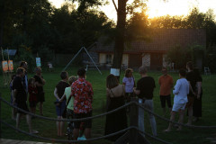 Grupa osób stoi w półkolu o zachodzie słońca. W głębi kadru zabudowania. Na pierwszym planie dwa sznury ogrodznia.
