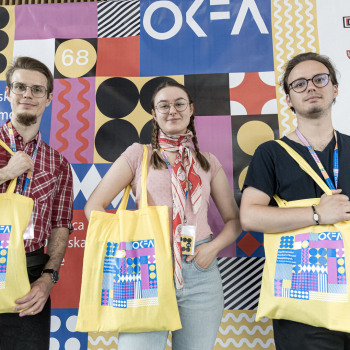 Dwóch chłopaków i dziewczyna stoją na tle banaru OKFA i pozują do zdjęcia z przewieszonymi przez ramię żółtymi torbami z logotypem OKFA.