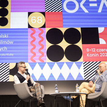 Na scenie zasiadający w szarych  fotelach Łukasz Maciejewski i Krzysztof Pągowski. W tle wyświetlony slajd OKFA.
