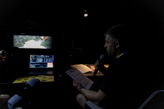 Mrok. Po prawej Bartek Nowakowski z CKiS w Koninie siedzi w fotelu. W prawej dłoni trzyma mikrofon, w lewej podkładkę z kartkami. Po lewej monitory.