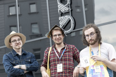 Trzech uczestników OKFA przed budynkiem CKiS-DK Oskard. Od lewej: chłopak w kapeluszu, chłopak z katalogiem na głowie oraz trzeci - prezentujący identyfikator.