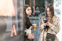 Lewa część karu odbicie szklanych drzwi. Po prawej dwie dziewczyny. Jedna z nich trzyma kubek ze słomką i patrzy w obiektyw. Druga patrzy w wyświetlacz telefonu.