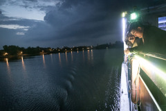 Noc. Dwie osoby opierają się o burtę statku. Po lewej tafla jeziora i brzeg oświetlony latarniami.