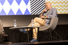 Andrzej Pągowski siedzący w fotelu na scenie. Przed nim stolik, a na nim dwie butelki z wodą. Po lewej nogi w kraciastych spodniach.
