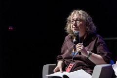 Maria Zmarz-Koczanowicz w fotelu. Na kolanach rozłożone kartki, w lewej dłoni mikrofon. Za nia mrok. W lewym górnym rogu dwa czerwone punkciki.