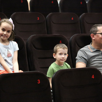 W czarnych fotelach kinowych z czerwoną numeracją siedzą od lewej: dziewczynka, chłopiec i śmiejący się mężczyzna. Dzieci patrzą wprost w obiektyw.