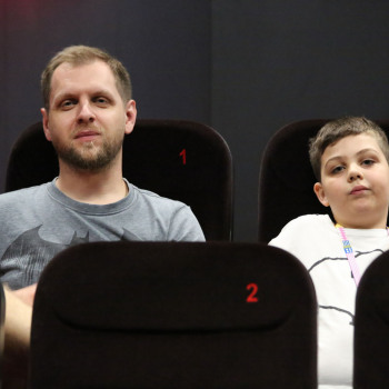 W fotelach kinowych siedzą: mężczyzna (z numerem 1) i chłopiec (z numerem 2). Patrzą w obiektyw.