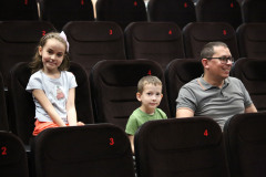 W czarnych fotelach kinowych z czerwoną numeracją siedzą od lewej: dziewczynka, chłopiec i śmiejący się mężczyzna. Dzieci patrzą wprost w obiektyw.