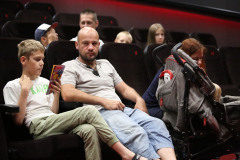 Publiczność w sali kinowej. W pierwszym rzędzie siedzą od lewej: chłopiec przeglądający folder, mężczyzna i kobieta z wózkiem.