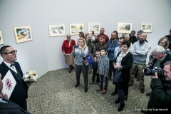 Robert Brzęcki, pod ostrzałem fotografów, przemawia do publiczności zgromadzonej w galerii. W prawej dłoni trzyma katalog z hasłem Sztuka nas Szuka, w lewej - bukiet kwiatów