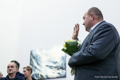 Sfotografowany z żabiej perspektywy Tomasz Olszewski podnosi do góry prawą rękę.  W lewym dolnym rogu widoczna mała grupa widzów