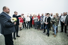 Zdjęcie grupowe. Po lewej: Tomasz Olszewski z bukietem kwiatów, obok kurator Robert Brzęcki wskazuje coś lewą ręką. Większość  osób patrzy we wskazywanym przez niego kierunku