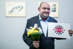 Tomasz Olszewski pozuje do zdjęcia. W prawej dłoni trzyma bukiet kwiatów (żólta, białą i zielona kompozycja), w lewej kartkę formatu A4 z napisem Sztuka nas Szuka