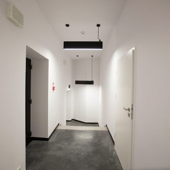Wąski korytarz o białych ścianach i szarej podłodze. Z sufitu zwisają dwie czarne prostokątne lampy .