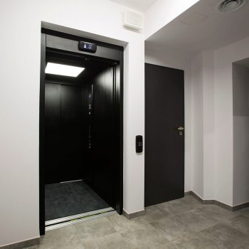 Otwarte drzwi windy osobowej. Po prawej para ciemnych drzwi.