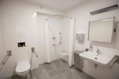 Łazienka przystosowana do potrzeb niepełnosprawnych. Po lewej muszla, na środku prysznic z zasłonkami, po prawej umywalka. Wnętrze utrzymane w jasnej kolorystyce.