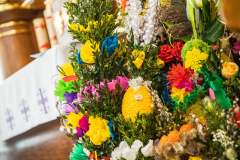 Gęsto zdobiona palma wielkanocna wykonana z kolorowych kwiatów oraz rozmaitych dekoracji, takich jak jajka, bazie, gałązki.