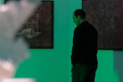 Odwrócony bokiem ubrany na ciemno mężczyzna ogląda wystawę. Ściany, na których wiszą prace oświetlone zielonym swiatłem.