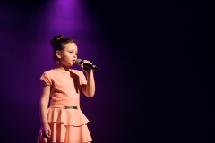 Dziewczynka uczesana w kok, w łososiwoej sukience z falbanami. W lewej ręce trzyma mikrofon.