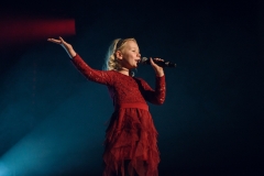 Dziewczynka w czerwonej sukience z falbankami z tiulu. W lewej ręce trzyma mikrofon, lewą wznosi w górę.