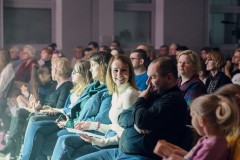 Publiczność oglądająca występy. Na piewrszym planie uśmiechnięta kobieta w białej bluzce, obok niej siedzi mężczyzna trzymający palec na ustach.