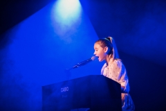 Dziewczyna przy keyboardzie śpiewa do mikrofonu. Niebieskie tło.
