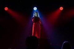 Oświelona na czerwono dziewczyna śpiewa do mikrofonu, który trzyma w prawej dłoni. Trzy reflektory: czerwone po bokach, pośrodku biały.
