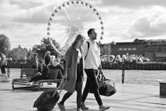 Kobieta ciagnie walizkę na kółkach. Obok niej mężczyzna z torbą podróżną. W tle rzeka, karuzela  i napis Gdańsk. Zdjęcie czarno-białe.