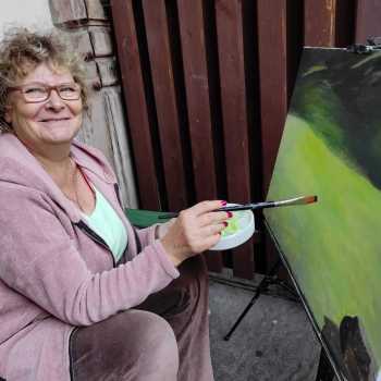 Uśmiechnięta kobieta pozuje przy swoim obrazie. Praca przedstawia pejzaż. W prawej ręce trzyma pędzel, w lewej tackę.