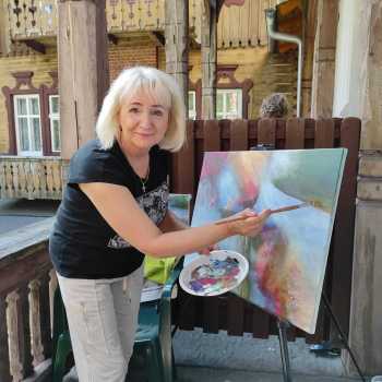 Kobieta na balkonie podczas malowania obrazu pozuje do zdjęcia. W tle fragment budynku w stylu tyrolskim.