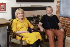 Danuta i Henryk Drzewieccy siedzący w fotelach. Uśmiechają się do zdjęcia. Kobieta ubrana w żółtą sukienkę, w głoni trzyma ciemną filiżankę na spodku. Mężczyzna w ciemnej koszulce i brązowych spodniach na szelkach.