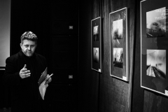 Marek Lapis spogląda w obiektyw.  Stoi obok zawieszonych na tle czarnej tkaniny fotografii.
