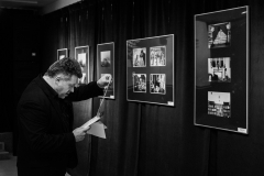 Marek Lapis.  Stoi obok zawieszonych na tle czarnej tkaniny fotografii. Czyta dokument.