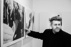 Marek Lapis wskazuje palcem prawej dłoni na zdjęcie zawieszone pod sufitem. Przedstawia ono jednego z braci Kaczyńskich i kobietę.