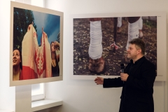 Marek Lapis trzyma mikrofon.  Patrzy na jedną z dwóch fotografii zawieszonych pod sufitem. Przedstawia trzymaną przed dwie kobiety flagę Polski. Po prawej zdjęcie owiniętych białym materiałem nadpęci konia.