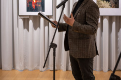 Filip Kowalkowski sfotografowany z lewego profilu przy mikrofonie. W prawej ręce trzyma kartki, lewą gestykuluje. Ubrany w ciemne dżinsy i marynarkę w kratę. Za nim prace zawieszone na jasnej kotarze.