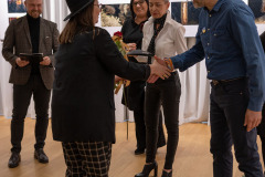 Zdjęcie grupowe. Na pierwszym planie Paweł Kosicki ściska dłoń kobiety w czarnym kapeluszu, czarnej koszuli i spodniach w kratę.