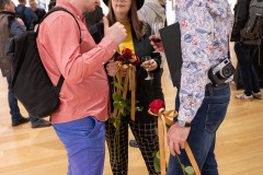 Dwóch mężczyzn i kobieta w trakcie rozmowy. Wszyscy trzymają czerwone długie róże ozdobione pomarańczowymi wstążkami.