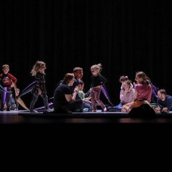 Grupa dorosłych i dzieci na scenie. Siedzą lub stoją. Za nimi sceniczny mrok. Zdjęcie zrobione z perspektywy widowni.