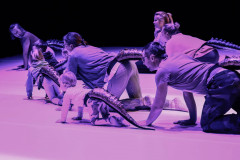 Grupa osób chodzi na czworaka po scenie. Mają przypięte macki ogony. Zdjęcie z użyciem kolorowego filtra.