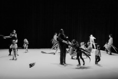 Dorośli i dzieci tańczą na scenie. Wszyscy mają przypięte ogony. Zdjęcie czarno-białe.
