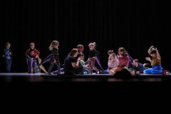 Grupa dorosłych i dzieci na scenie. Siedzą lub stoją. Za nimi sceniczny mrok. Zdjęcie zrobione z perspektywy widowni.