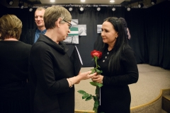 Ubrana na ciemno, krótko ostrzyżona kobieta z katalogiem rozmawia z Izą Kosiukow. Artystka trzyma czerwoną różę. Za nimi sylwetki rozmawiających i oglądających prace